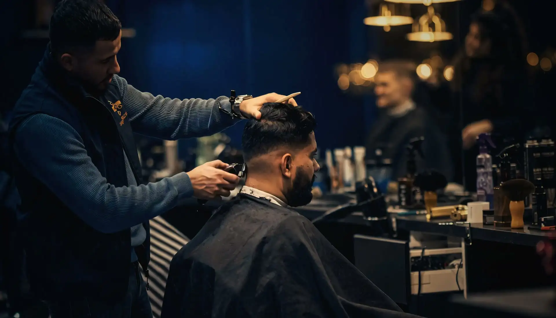 Barberare klipper en fade i en barbershop med mässingslampor och mörkblå vägg